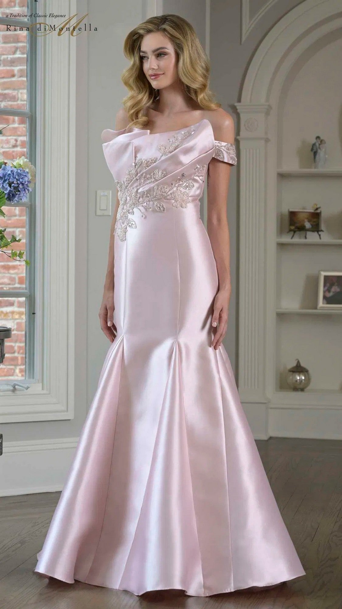 A woman stands indoors wearing the Rina Di Montella RD3408, a blush long formal dress with an off-the-shoulder neckline, floor-length satin skirt, and floral embellished bodice.